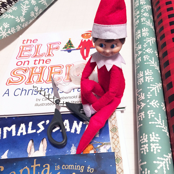 Elf on the Shelf sitting atop kids Christmas books with a pair of scissors and wrapping paper