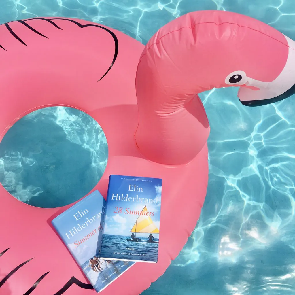 Inflatable pink flamingo floating in a pool with two Elin Hilderbrand books sitting on the pool float. The books are Summer of 69 and 28 Summers