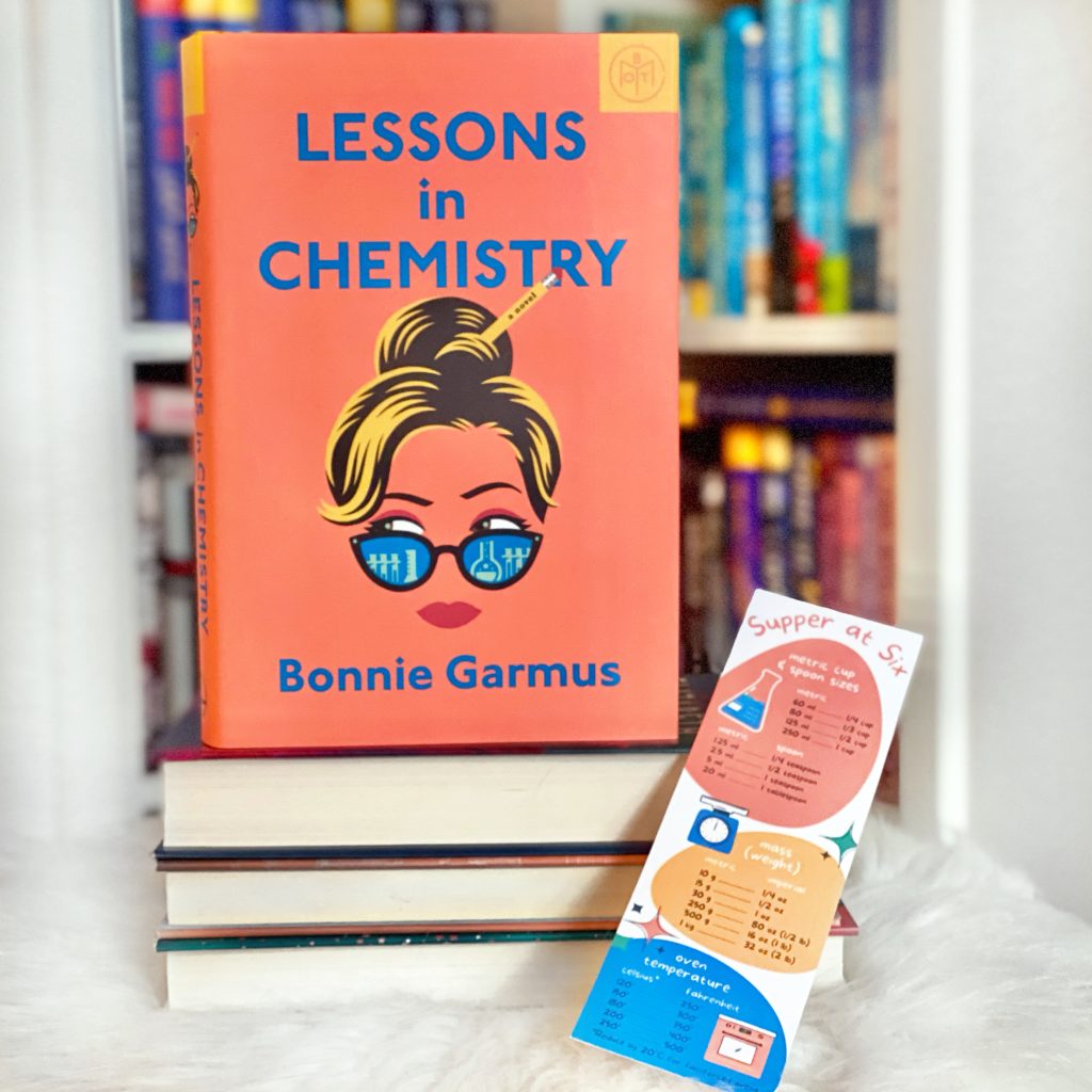 The book Lessons in Chemistry standing on top of a stack of other books. Leaning against the books is a bookmark in coordinating colors with a list of kitchen conversions.