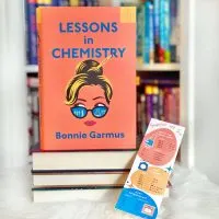 The book Lessons in Chemistry standing on top of a stack of other books. Leaning against the books is a bookmark in coordinating colors with a list of kitchen conversions.
