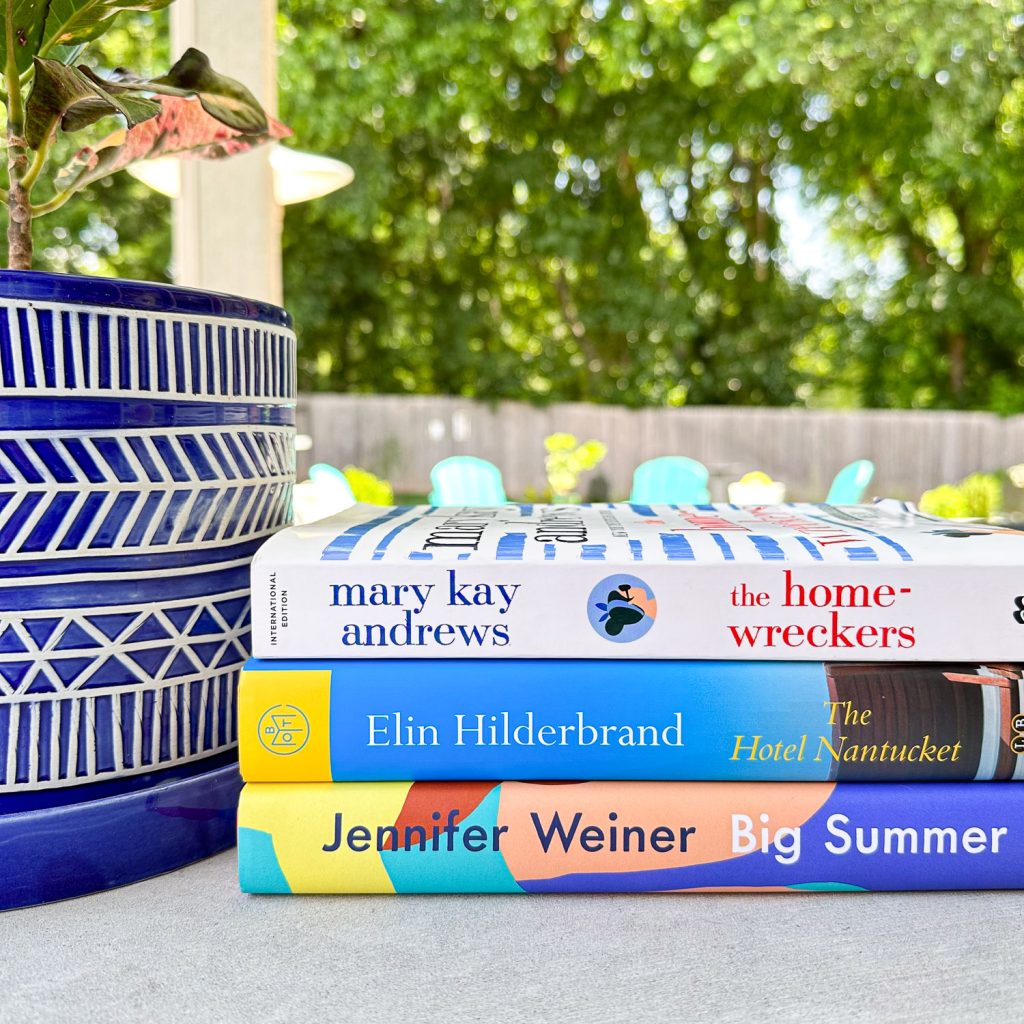 A stack of three books sitting on a patio table featuring authors similar to Mary Kay Andrews. The books are Big Summer by Jennifer Weiner, The Hotel Nantucket by Elin Hilderbrand, and The Homewreckers by Mary Kay Andrews.  
