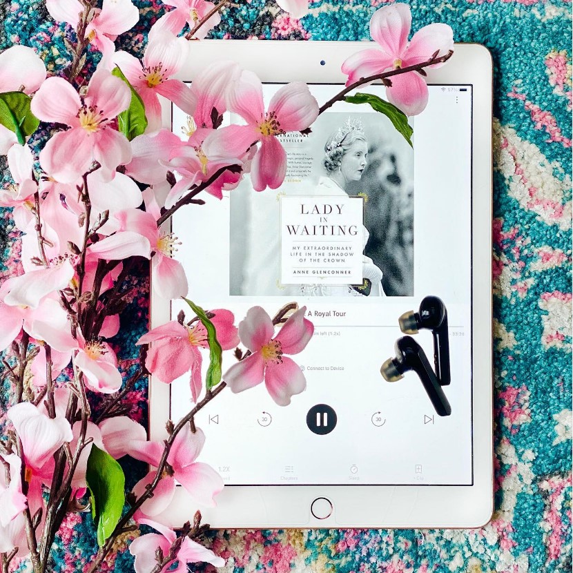 Flatlay photo of the audiobook version of Ann Glenconner's memoir Lady in Waiting open on an iPad screen. The iPad is sitting ona teal and pink rug with pink flowers overlapping the iPad and a pair of black earbuds rest on the iPad screen.