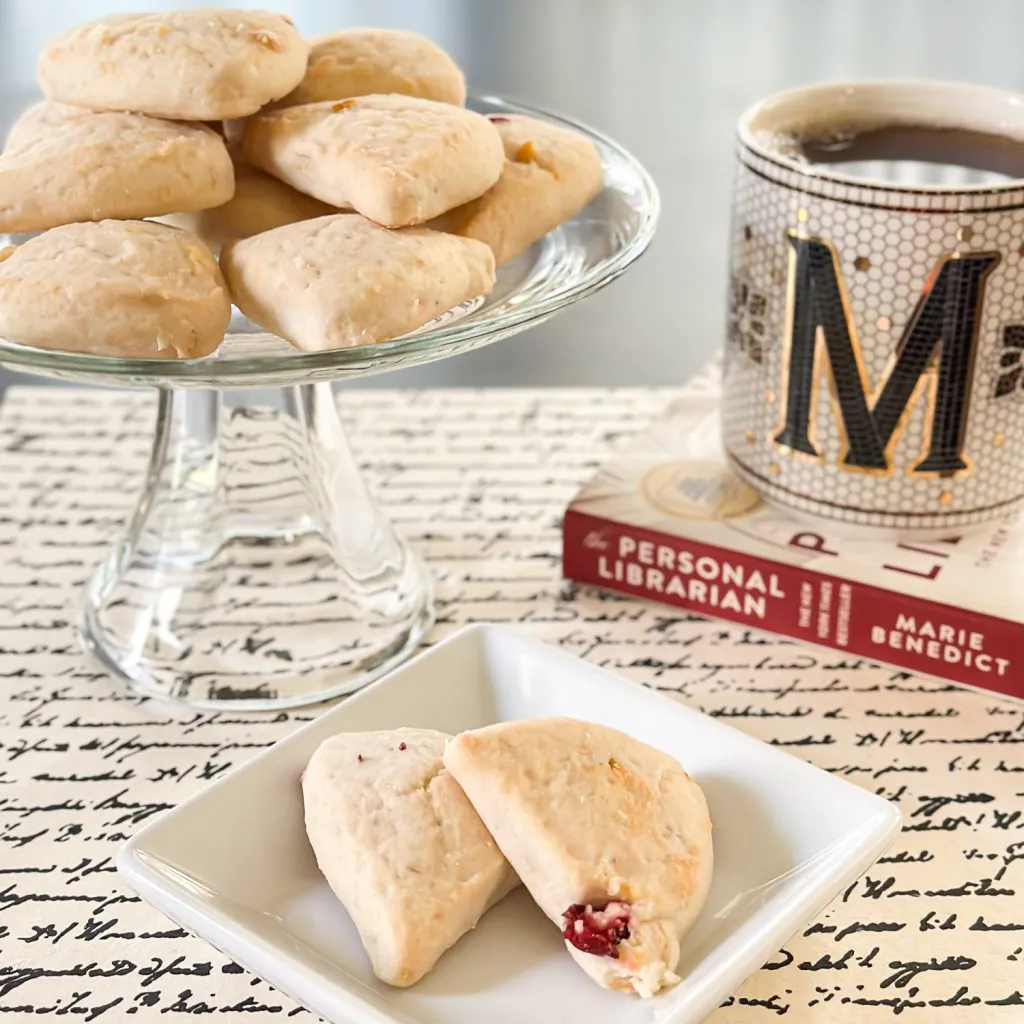 Two small scones on a white plate in front of a stand of scones and cup of tea on top of The Personal Librarian novel 