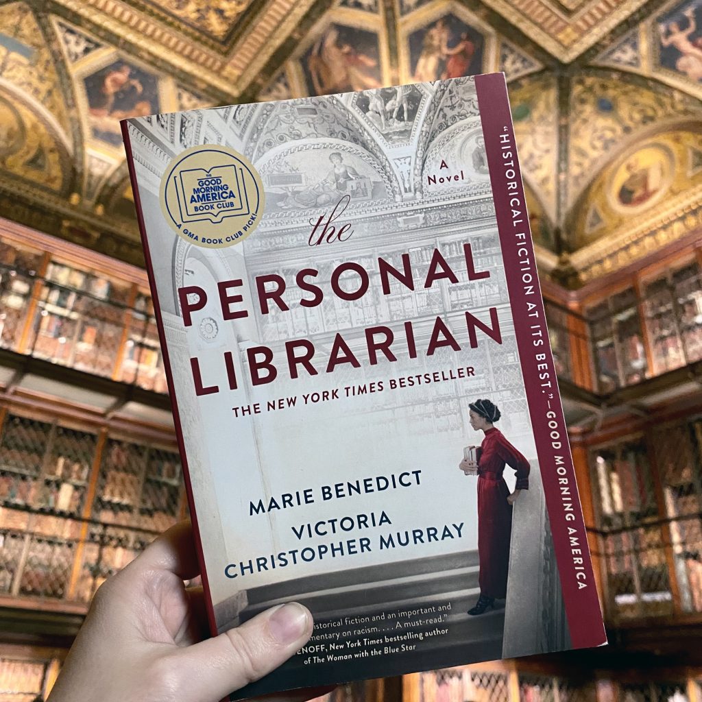 Paperback copy of The Personal Librarian in the foreground with the bookshelves and ornate ceiling of The Morgan Library in the background