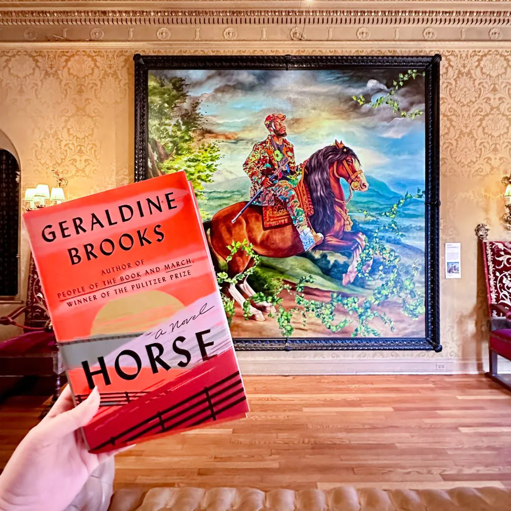 hand holding the book Horse by Geraldine Brooks in front of a painting by Kehinde WIley. 