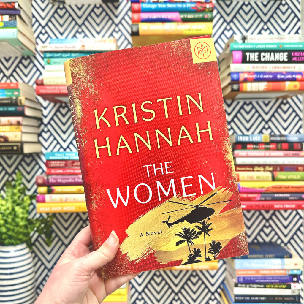 hand holding The Women by Kristin Hannah in front of a wall of books