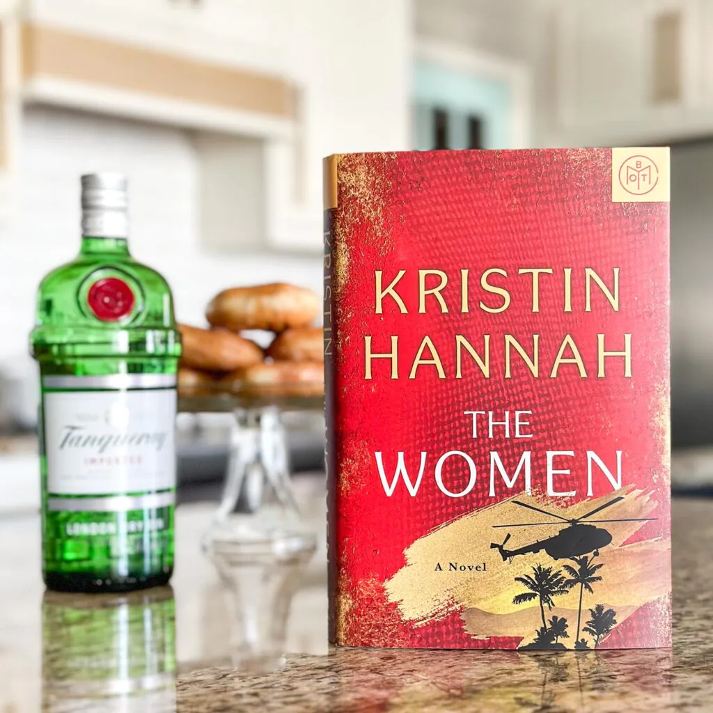 PHoto of the novel The Women sitting on a counter next to a bottle of gin and tray of donuts