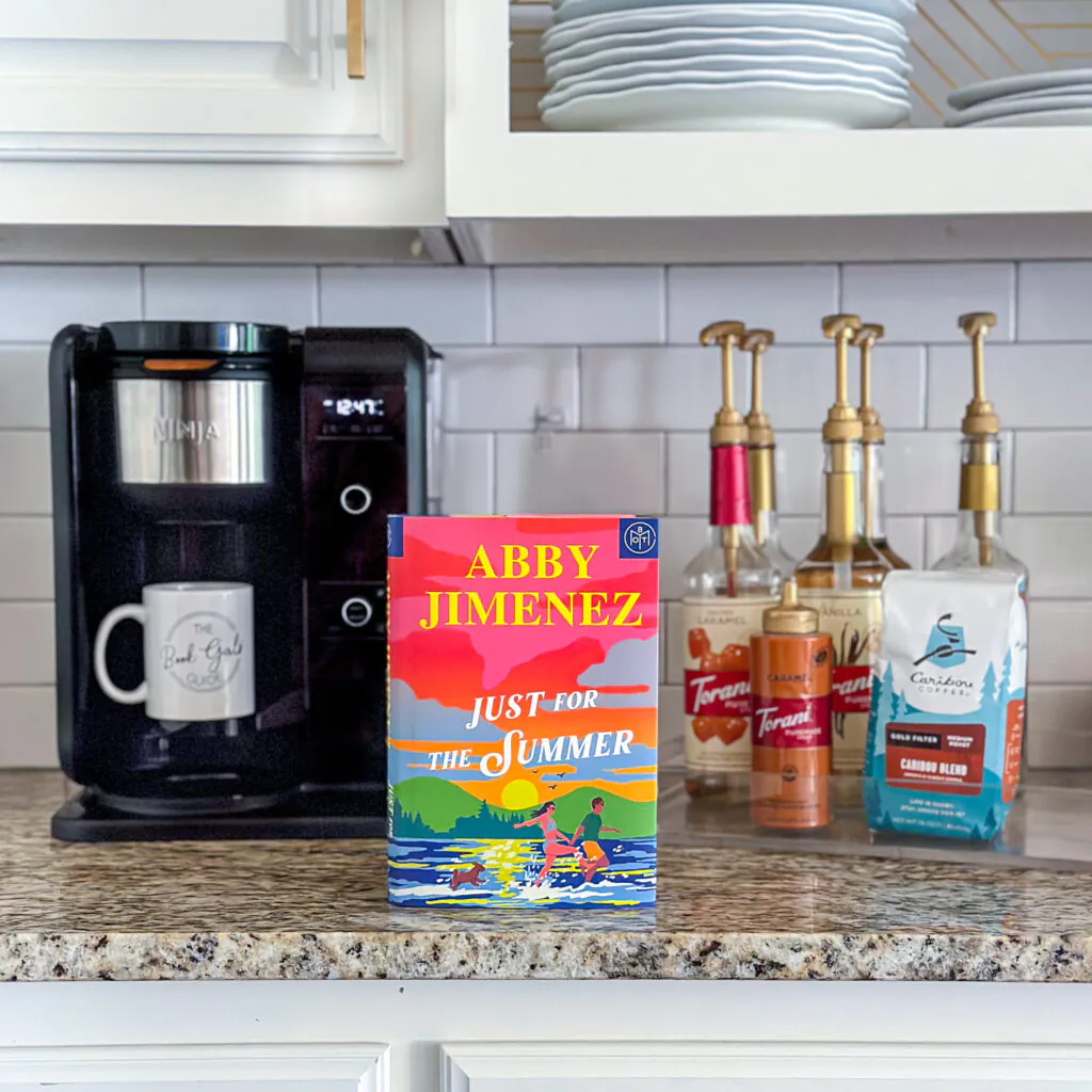 Hardback copy of Just for the Summer by Abby Jimenez sitting on a counter by a coffee maker and flavored coffee syrups