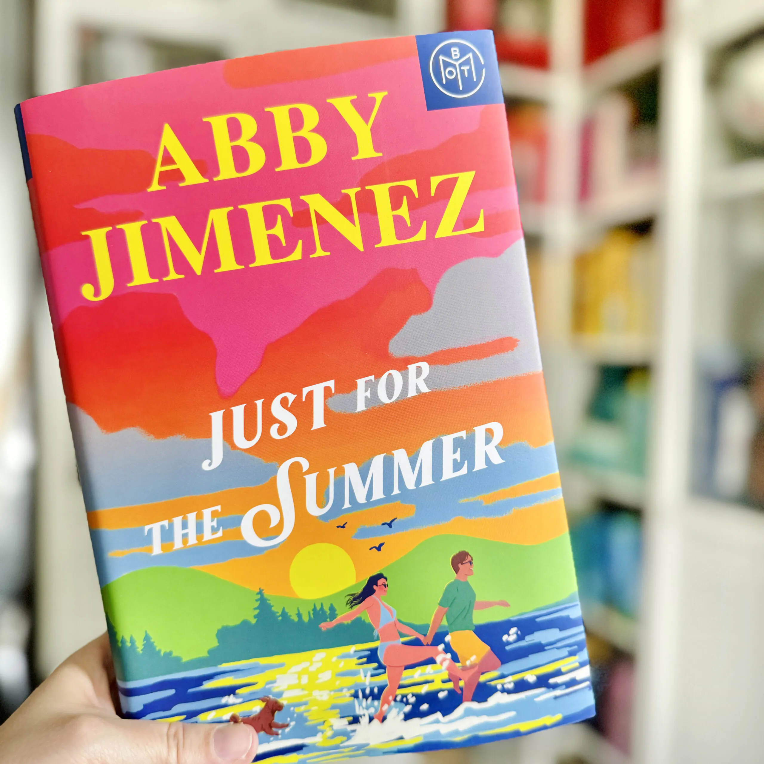 A hand holding a copy of the book Just for the Summer by Abby Jimenez in front of a white bookshelf with books organized by color