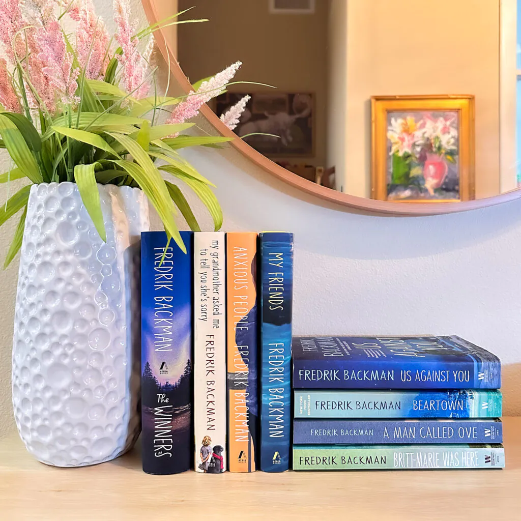 A photo of all eight of Fredrik Backman's full length novels sitting on a table next to a vase of flowers and below a round mirror