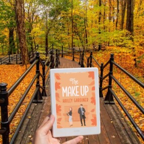Photo of a wooden walkway surrounded by trees and fall leaves and a hand holding out a Kindle displaying a fall book cover