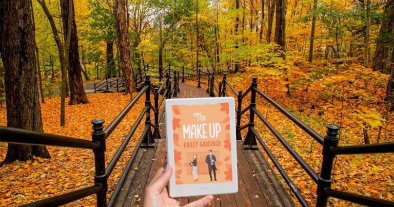 Photo of a wooden walkway surrounded by trees and fall leaves and a hand holding out a Kindle displaying a fall book cover