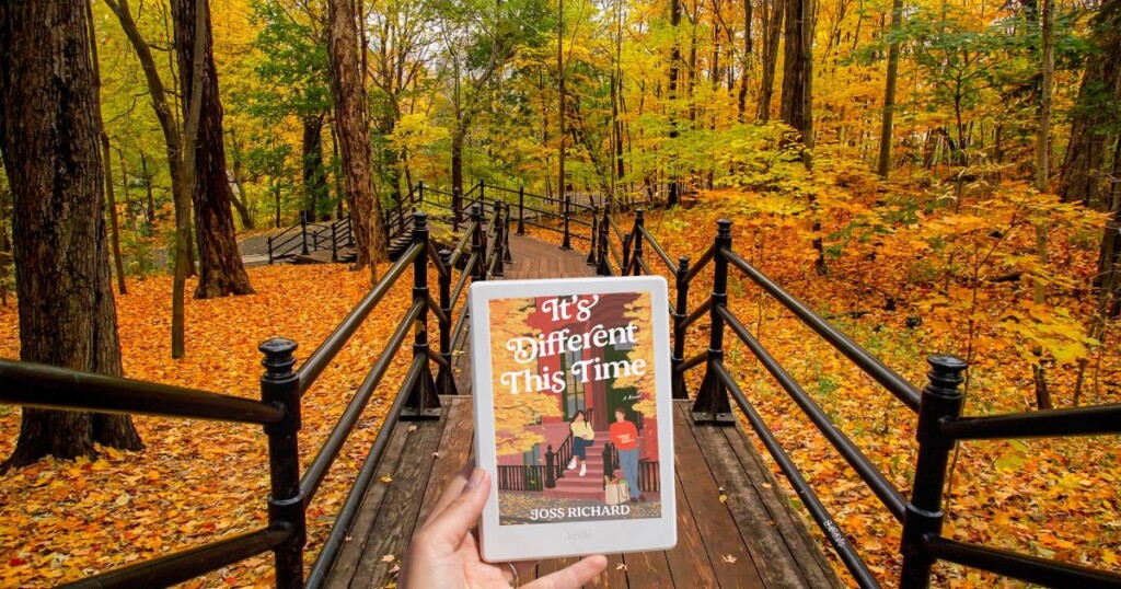 Photo of a wooden walkway surrounded by trees and fall leaves and a hand holding out a Kindle displaying a fall book cover