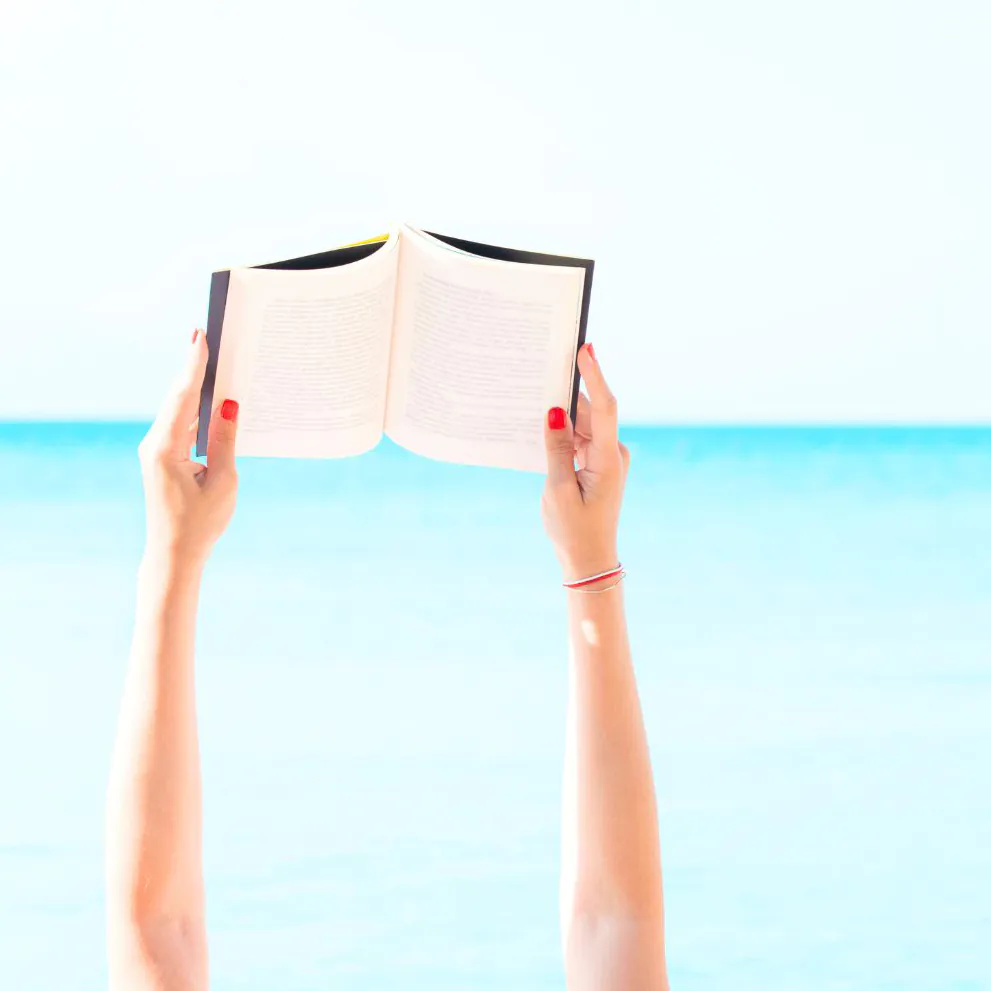 Arms holding an open book high in the air with the ocean in the background