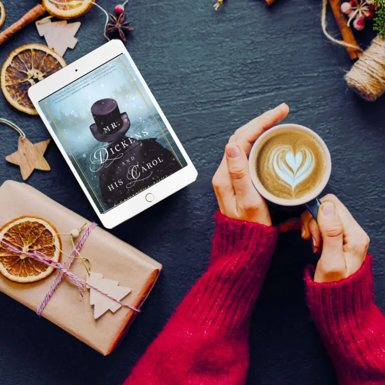 Hands holding a mug with a latte on a table covered in brown paper wrapped gifts and an iPad with the cover of Mr. Dickens and His Carol on the screen
