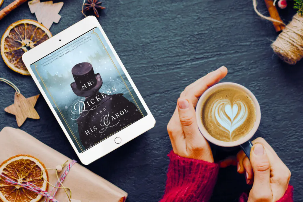 Hands holding a mug with a latte on a table covered in brown paper wrapped gifts and an iPad with the cover of Mr. Dickens and His Carol on the screen