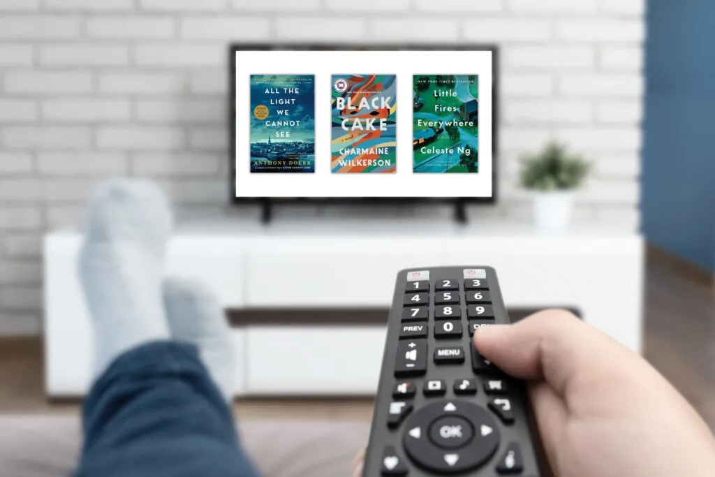 A hand holding a remote and feet up on an ottoman in the foreground with a TV screen in the background that has three book covers on the screen